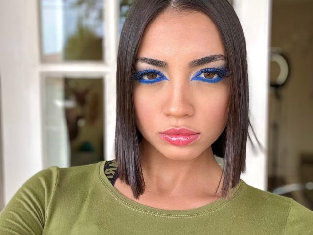 CameliaDeihl has blue eyeliner, glossy pink lips, and a medium green top, with straight dark brown bob haircut, brown eyes, and a serious expression.