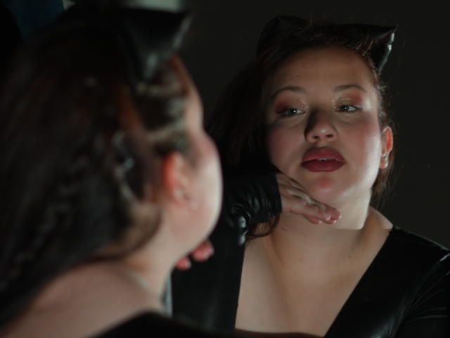 RavenBlaze with cat ears and black gloves, face slightly red, looking at her reflection, dark brown hair, red lipstick, black top, hand near chin, soft light, dark background.
