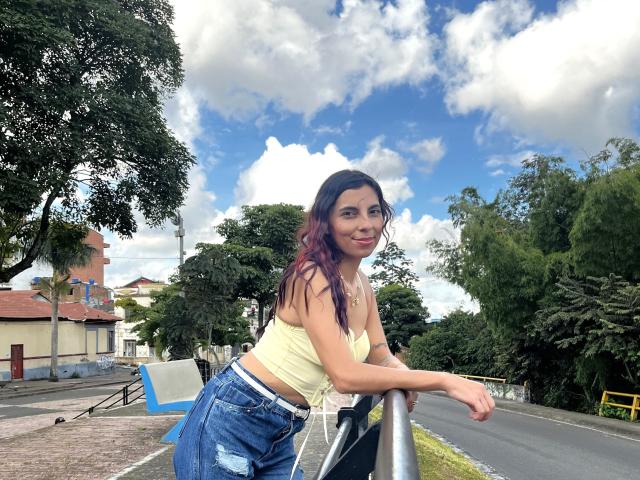 CarlaTorres with long purple hair, yellow tube top, and blue jeans, leaning on a bike rack, smiling, outdoors, under a blue sky with fluffy clouds.
