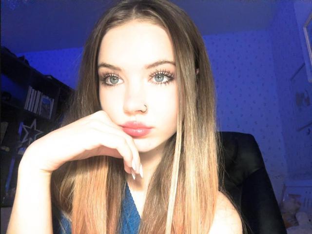 FannyEnsell has long brown hair, green eyes, and pink lipstick. She's looking at the camera with her hand near her face, wearing a blue top. She has a nose ring.