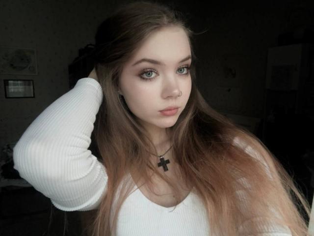FannyEnsell with long brown hair, white ribbed top, cross necklace, green eyes, and nose ring, hand in hair, serious expression, light skin, dark room.