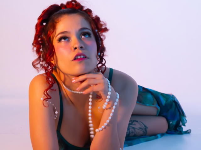 ArielleBrooks with curly red hair, green top, pearl necklace, and tattoo, lying on white, looking up with hand on chin, glowing pink blush, blue-green skirt, and boots.