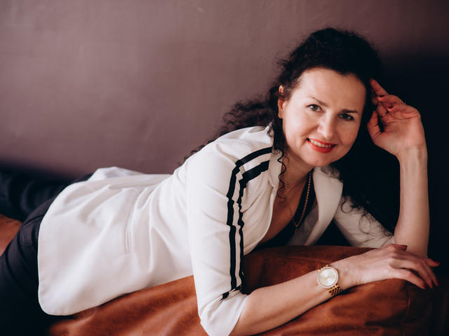 MiroslavaHappi lying on a brown surface, wearing a white shirt with black stripes, black pants, and a gold watch, smiling with curly black hair, red lipstick, and a necklace.