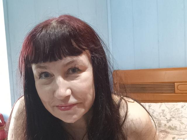 Mila69s ha capelli neri, lisci e con frangia, pelle chiara e piccoli seni. Sorride con il rossetto rosso, guardando nella fotocamera. Indossa un top senza spalline..