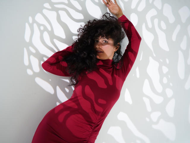 SugerVibes with curly black hair, red long-sleeve shirt, shadowed white wall, dark lipstick, looking to the side, sunlight casting leaf patterns.