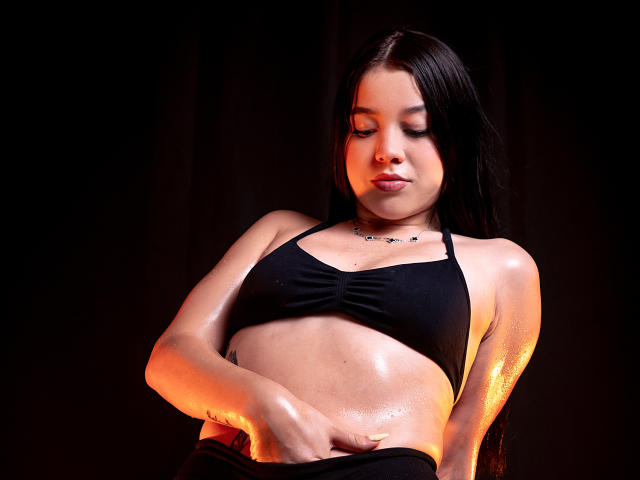 EmilyFrostt with shiny, sweaty skin, wearing a black sports bra, hand on her belly, glistening in orange light, dark background, long black hair, subtle necklace.