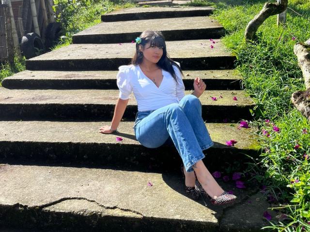 AshlyLee with dark hair, white blouse, blue jeans, and leopard shoes, sitting on stone steps, flowers in her hair, holding petals, grass and flowers around, sunny day.