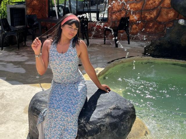 AshlyLee with tits in a blue floral dress, sitting on a rock by a green water feature, splashing water with her hand, wearing a pink headband, smiling.