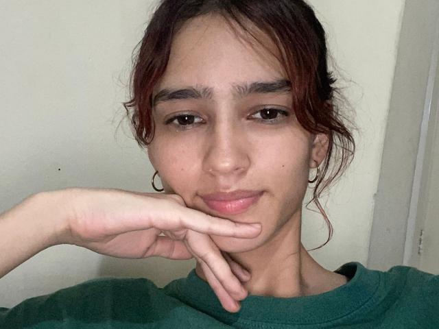 MelodyMansson has red hair, brown eyes, and is wearing a green shirt. She's resting her chin on her hand, showing a slight smile with pink lips.