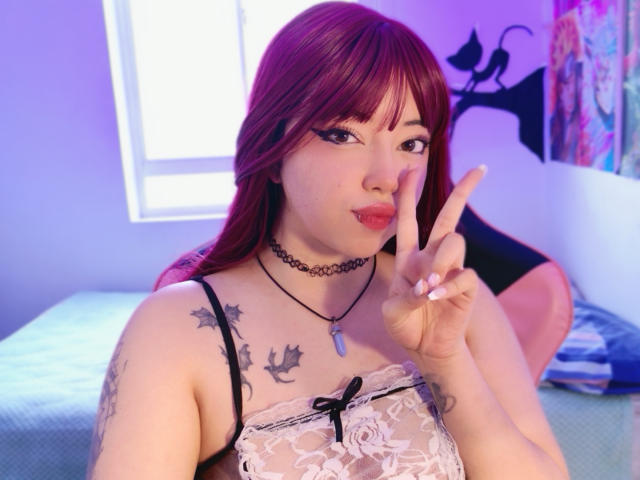 Arieyaara with red hair, black eyeliner, and a choker, showing a peace sign, wearing a lace top, revealing tattoos on her chest and arm, in a purple-lit room.