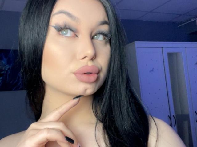 Rosell with long black hair, blue eyes, and pink lips, pouting, pointing with black-nail-ed finger, wearing black top, looking at camera, in blue-lit room.