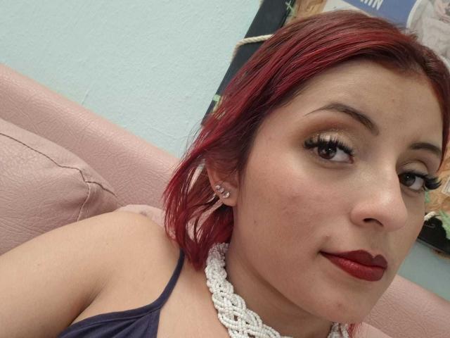 LizBonnet tiene el pelo rojo, piel clara y usa un lápiz labial rojo oscuro. Está mirando a la cámara con pestañas largas, y lleva un collar de encaje blanco y una blusa oscura..