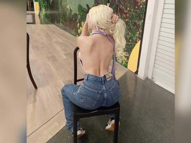 SabrinaColle, avec ses cheveux blonds, dos à la caméra, portant un haut violet à encolure dégagée, un jean bleu et des baskets, assise sur une chaise noire, exposant son dos nu et ses fesses..