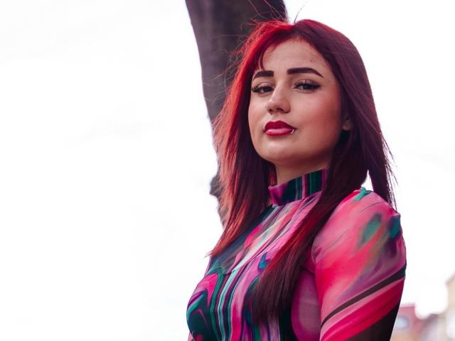 Julieta Dupont, met rood haar, draagt een kleurrijke, strakke top met een patroon, heeft rode lippenstift en een lichtelijk zelfvoldane uitdrukking, en staat met een boom op de achtergrond..