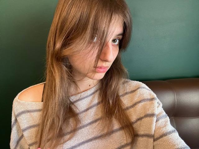 KarieMestler with long brown hair, striped beige sweater, and green wall, looking slightly to the side, with a subtle pout. Brown leather chair in the background.