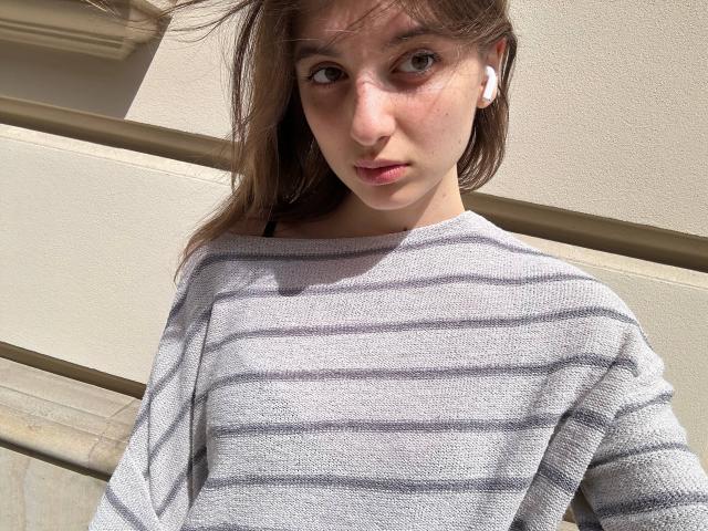KarieMestler with pale skin, brown eyes, and light brown hair, wearing a gray striped sweater, has white earbuds in, looking slightly annoyed. Sunlight casts shadows on her face.