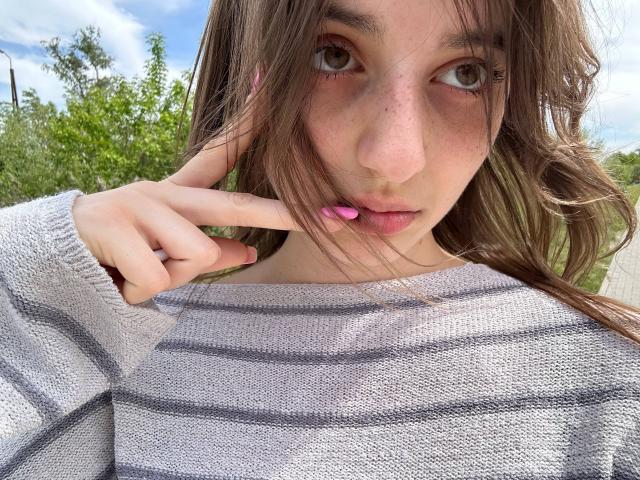 KarieMestler with pink lipstick, brown wavy hair, and freckles, wearing a gray striped sweater, touching her lips with a finger, looking into the camera.