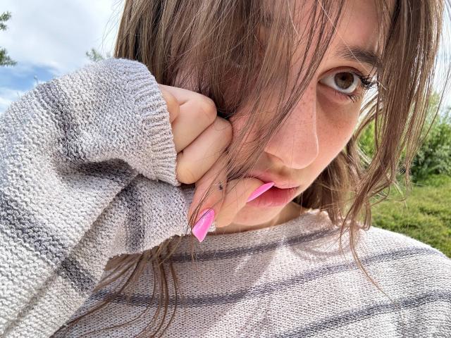KarieMestler with messy brown hair, pink nails, grey sweater, and hand near mouth, showing a cute, curious expression, outdoors on a sunny day.