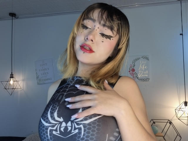 IvonnySakura, avec des cheveux blonds, un eye-liner noir, du rouge à lèvres rouge, et une robe avec un motif de toile d'araignée, touchant sa poitrine, les mains sur le cou, lumière douce, lampes suspendues, mur blanc, léger sourire..