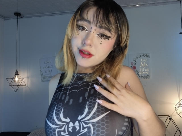IvonnySakura, con capelli che sfumano dal biondo al castano, eyeliner nero e rossetto rosso, indossa una maglietta nera con una ragnatela tatuata e tocca il petto, in una stanza poco illuminata..