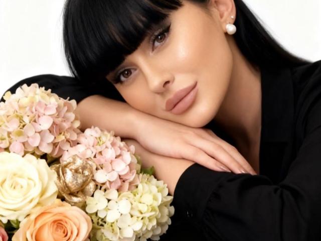NikkiLusty with black bangs, pearl earrings, and nude makeup, rests her chin on her hand, looking at the camera, with a bouquet of pink and white flowers.