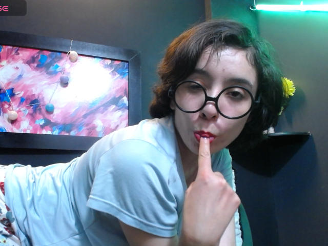 ElodiePerrine with short brown hair, glasses, and red lipstick, holding her finger to her lips, wearing a light blue shirt, and a floral patterned skirt. She's sucking on her finger.