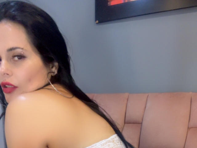 CharlotteCrofort with long black hair, red lipstick, and hoop earrings, looking over her shoulder with shiny bare shoulders, sitting on a pink upholstered seat.