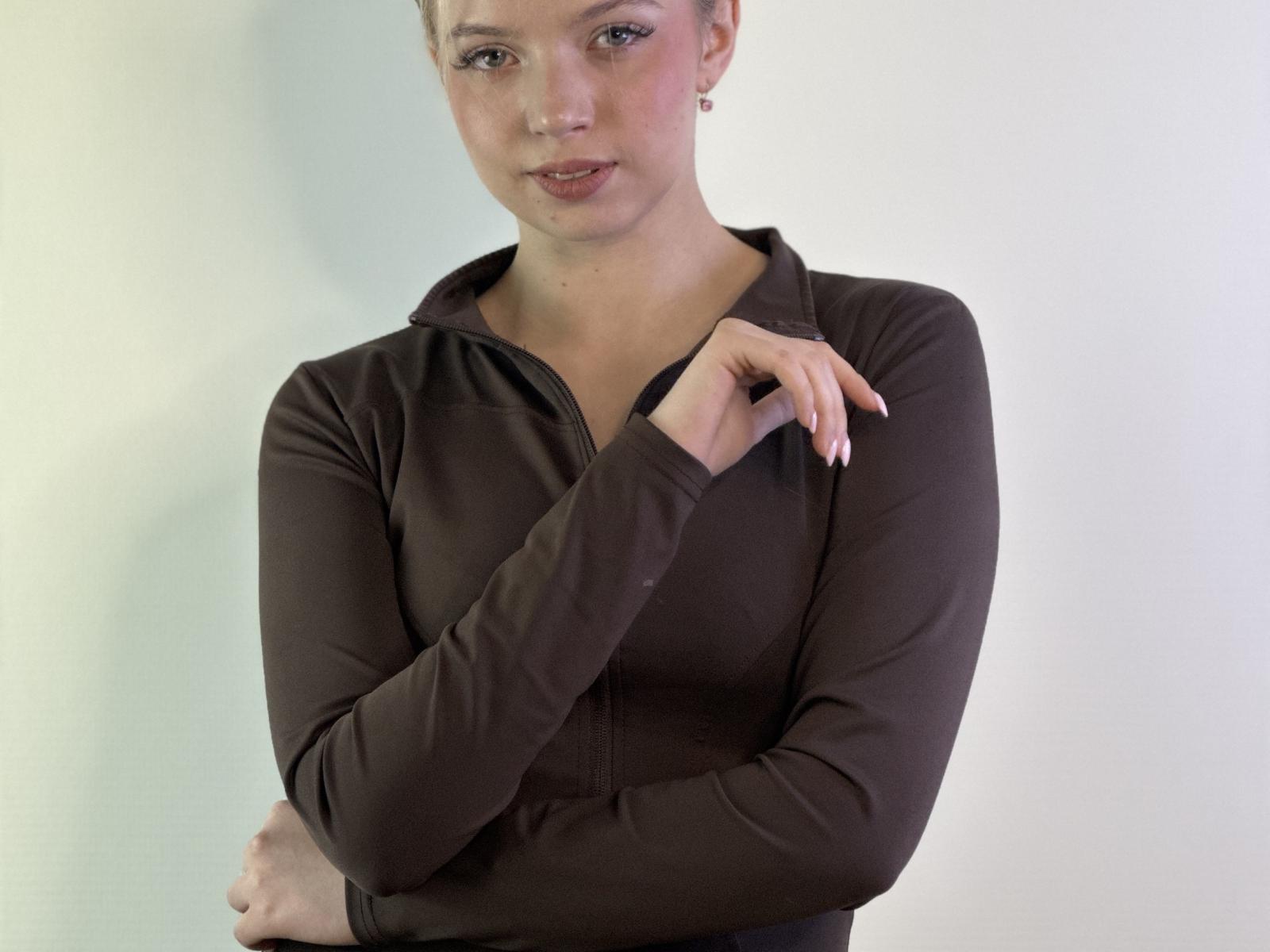 LitaHoltHouse heeft een lichte huid, bruine ogen en kort bruin haar. Ze draagt een bruin lang袖 top met een rits, en haar rechterhand is dicht bij haar borst, terwijl ze iets naar links kijkt..