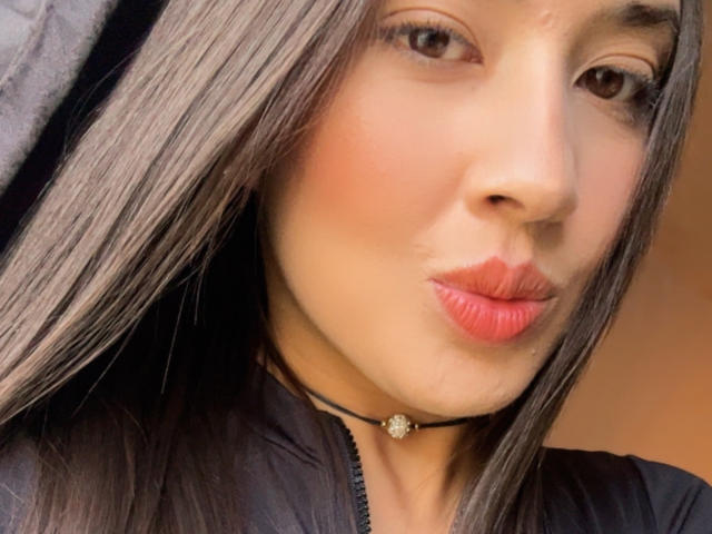EmmaLombardi's face with long, straight brown hair, wearing a black choker with a small gem, pouting pink lips, and a slight smile. Her brown eyes are centered.