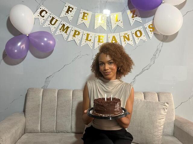 AylaCallowey with curly brown hair, tan skin, small tits, wearing a beige sleeveless top, sitting on a gray couch, holding a chocolate cake with candles. Balloons and banner in background.