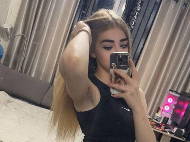 CarolannFettig with long blonde hair, black Nike tank top, arm raised, holding phone in mirror selfie, showing armpit and slight sideboob. Bedroom with bed and curtains.