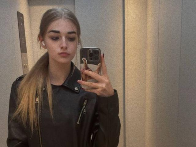 CarolannFettig with long blonde ponytail, black leather jacket, black top, holding phone, taking mirror selfie in elevator, serious expression, wearing earbuds, nails painted red.