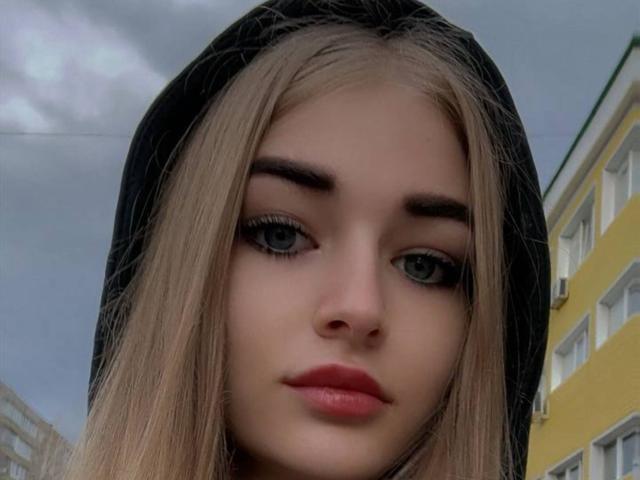 CarolannFettig has long blonde hair, blue eyes, and full pink lips. She's wearing a black hood, and her eyebrows are thick and dark. Her face is centered in the image.