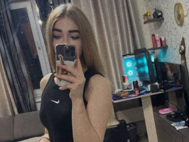 CarolannFettig with long blonde hair, wearing a black Nike sports bra, holding a phone in front of her face, showing her small boobs, in a cluttered room.