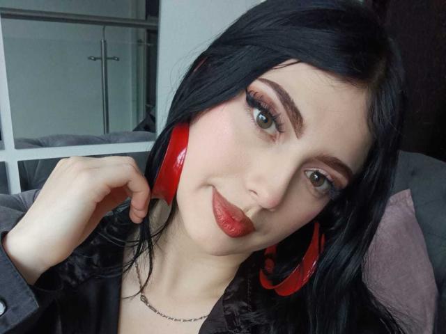 IsabellaaRossi with black hair, red headphones, and red lipstick, has green eyes and is touching her ear. She's wearing a black shirt and a necklace.