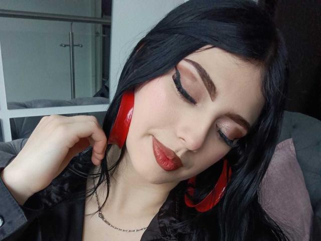 IsabellaaRossi with black hair, red headphones, closed eyes, and red lipstick, wearing a black shirt, hand near ear, looking relaxed.