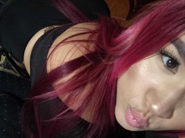 LucieCurie with pink hair, pouty lips, and dark makeup, wearing a black top, showing her shoulders and a tattoo on her upper arm. Her eyes are looking at the camera.