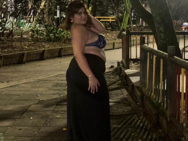 IvyOconner, standing on a sidewalk at night, wearing a blue lace bra and black skirt, showing her big ass and belly, with one hand in her hair.
