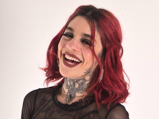 GabrielaTorres with red hair, smiling, dark lipstick, black lace top, tattooed neck, hoop earrings, looking happy, showing teeth, medium-length wavy hair, white background.