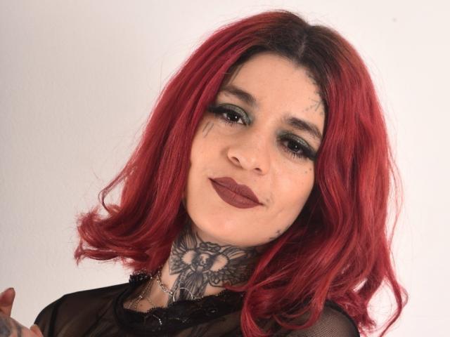 GabrielaTorres with red hair, dark lipstick, green eyeshadow, tattoos on neck, and a black sheer top, smiling slightly, showing a small nose piercing.