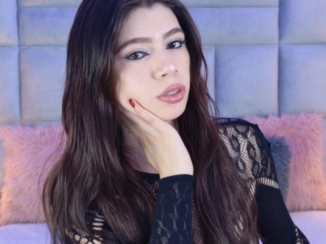 LindaMancini with long brown hair, wearing a black lace top, has red nails, slightly parted pink lips, and is touching her cheek with her right hand.