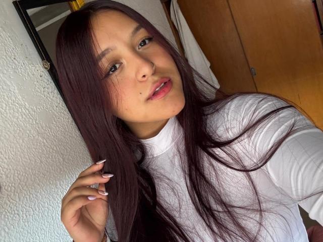 MarianaVargasX with long brown hair, white turtleneck, and pink lips, touching her hair, showing her small, cute nose and slightly visible tits, in a selfie.