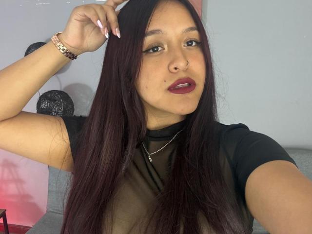 MarianaVargasX with long dark hair, red lipstick, black sheer top, nose ring, and pink bracelet, hand in hair, looking at camera, slight smile, showcasing her tits.