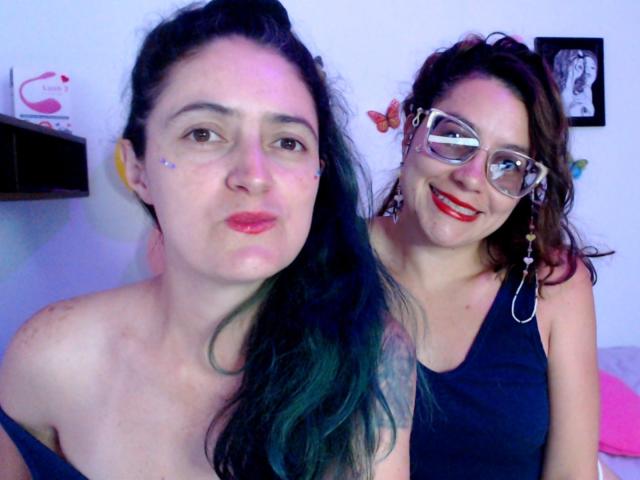 LunaVenus with dark green hair, no top, and small tits, next to LunaVenus with glasses, red lipstick, and black tank top, smiling in a bedroom.