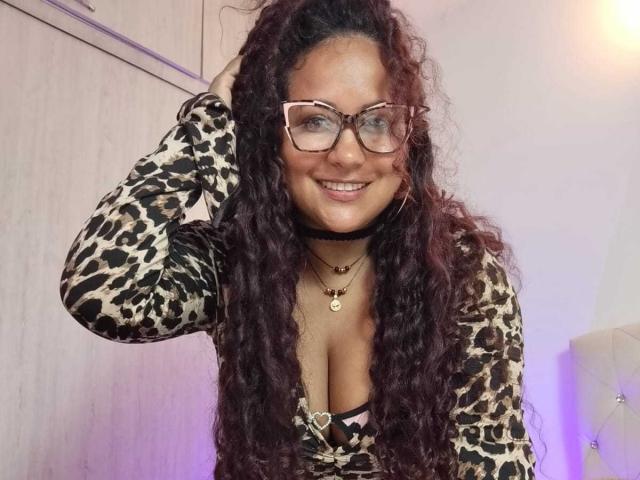 YiyaHot69, con cabello oscuro y rizado, usando gafas, una blusa de estampado de leopardo, un collar negro y un collar dorado, sonriendo, con la mano en el cabello, mostrando sus grandes senos. Fondo de dormitorio, luz púrpura..