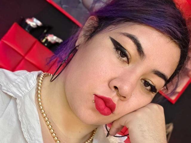 LilithVelvett with purple-dyed hair, black eyeliner wings, red lipstick, nose ring, gold necklace, pouty lips, resting cheek on hand, white shirt, and a serious expression.