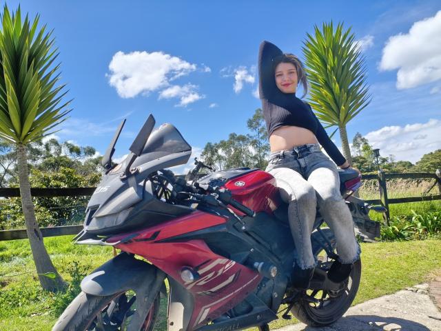 DarckVelvet, con pechos pequeños, sentada en una motocicleta roja, vistiendo una blusa negra, pantalones blancos y botas negras, bajo un cielo azul con nubes, y dos palmeras..