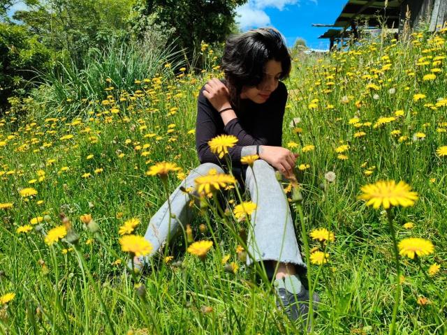 DarckVelvet, sentada en un campo de margaritas amarillas, tiene el pelo corto y negro, una camisa negra de manga larga y pantalones grises, con una expresión triste y pensativa, bajo un cielo azul brillante..
