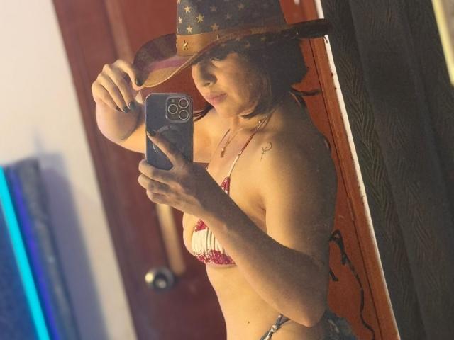 AnastasiaEvansX in einem roten und weißen Bikini, mit einem Cowboyhut, die ein Selfie mit einem Handy macht, wobei ihre Brüste und ein kleines Tattoo an ihrer rechten Schulter zu sehen sind..