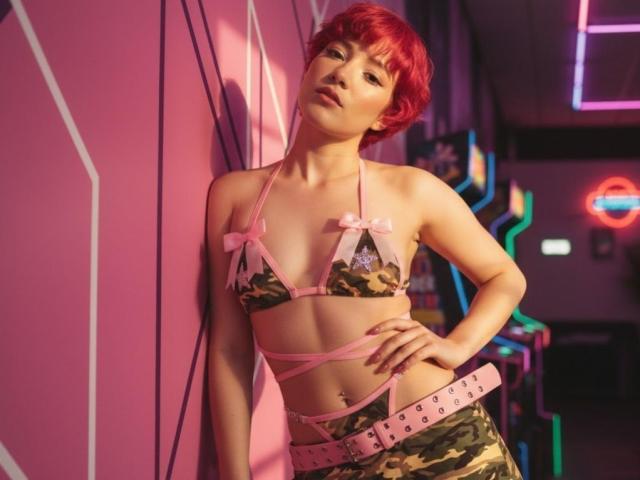MiaSinclairX with red hair, wearing a camo bikini top with pink bows and a pink belt, leaning against a pink wall in a neon-lit room. Boobs visible.